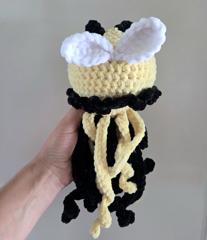 May include: A handmade crochet plush toy featuring a round yellow head with two white fuzzy ears, a black collar, and dangling black and yellow yarn tentacles. The soft, textured yarn construction creates a tactile surface with distinct color blocks.