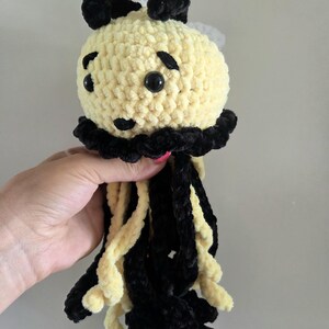 May include: A crocheted plush toy shaped like a jellyfish with a round, pale yellow head, black yarn accents, and two black ears on top. It features black plastic eyes, a small black nose, and long, dangling tentacles in alternating black and pale yellow.