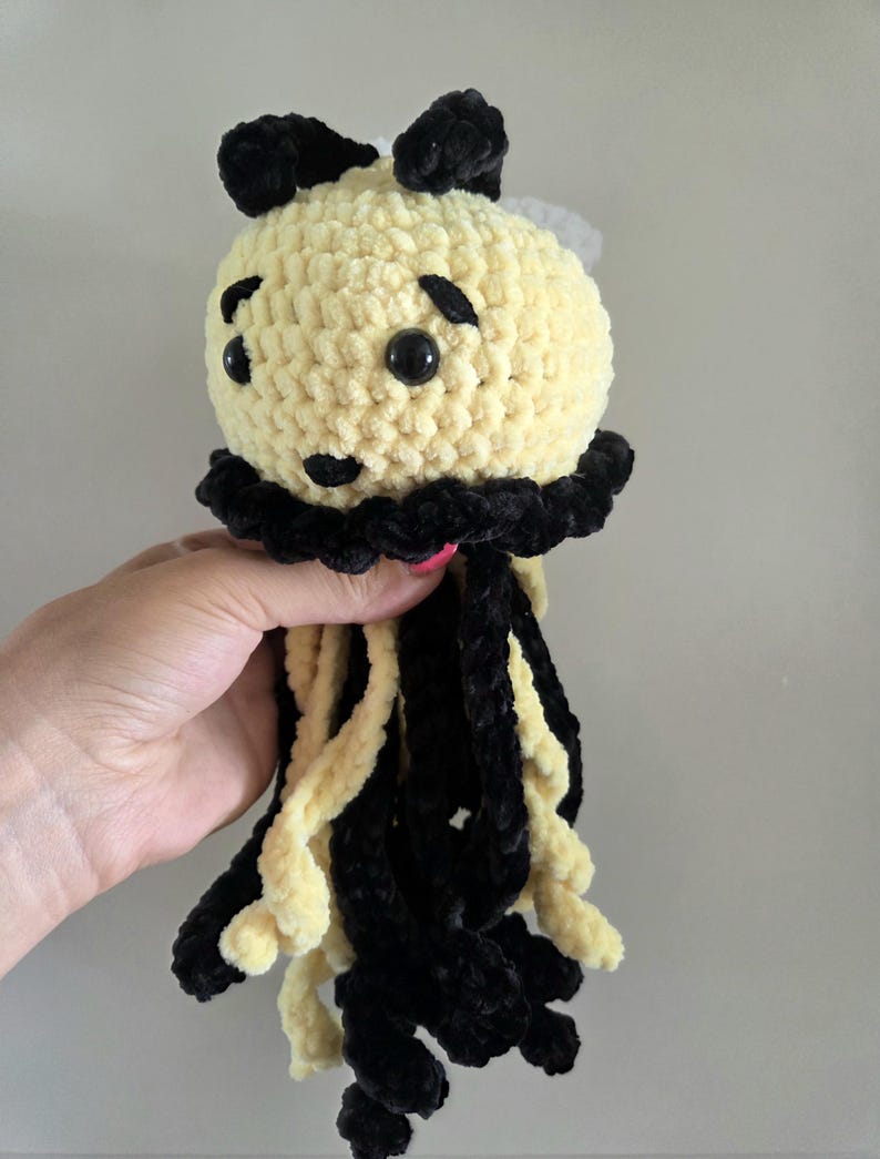 May include: A crocheted plush toy shaped like a jellyfish with a round, pale yellow head, black yarn accents, and two black ears on top. It features black plastic eyes, a small black nose, and long, dangling tentacles in alternating black and pale yellow.