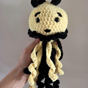 May include: A crochet jellyfish plush toy featuring a round, pale yellow head with black embroidered eyebrows, a small black nose, and two black plastic eyes. It has a black ruffled collar and long, curly tentacles in alternating black and yellow.