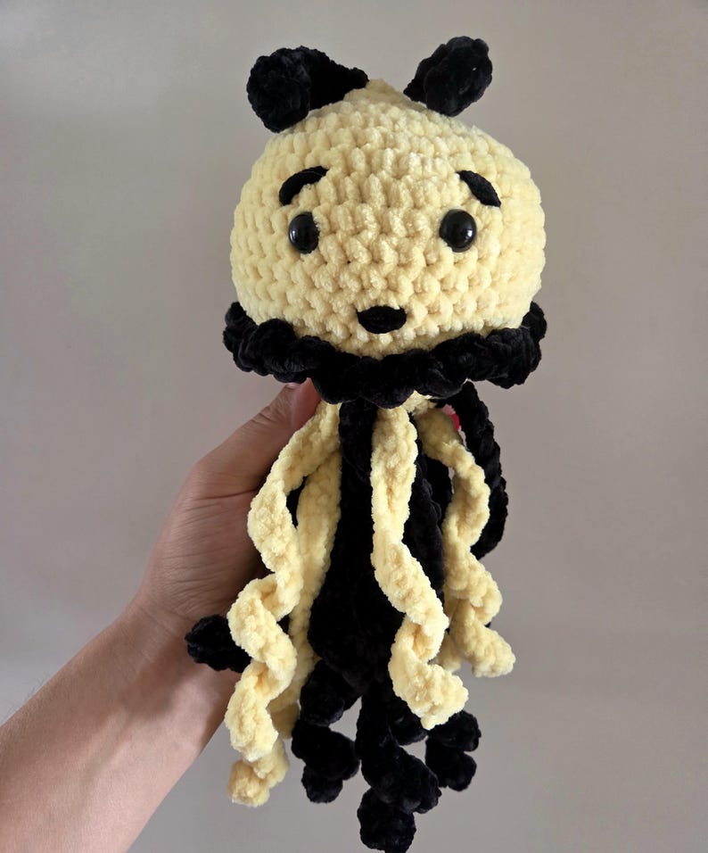 May include: A crochet jellyfish plush toy featuring a round, pale yellow head with black embroidered eyebrows, a small black nose, and two black plastic eyes. It has a black ruffled collar and long, curly tentacles in alternating black and yellow.