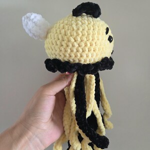 May include: A crocheted jellyfish plush toy featuring a round pale yellow head with a black top accent, a single white wing-like protrusion, and multiple dangling tentacles in alternating black and pale yellow yarn with a textured finish.