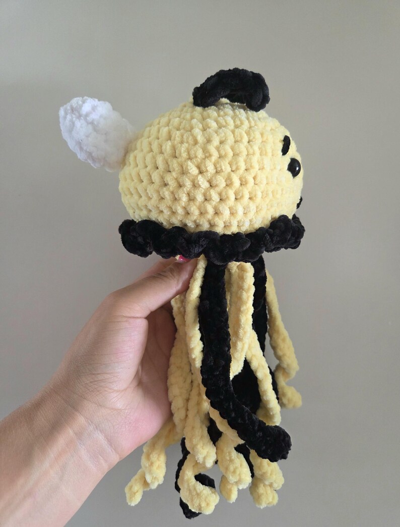 May include: A crocheted jellyfish plush toy featuring a round pale yellow head with a black top accent, a single white wing-like protrusion, and multiple dangling tentacles in alternating black and pale yellow yarn with a textured finish.