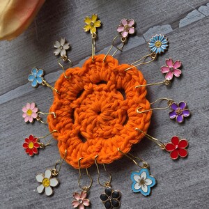 Peut inclure: Un cercle orange crocheté avec des marqueurs de point en forme de fleurs colorées. Les marqueurs de point ont des crochets dorés et sont de différentes couleurs, dont le jaune, le rose, le bleu, le rouge, le blanc et le violet. Le fond est une surface en bois grise.