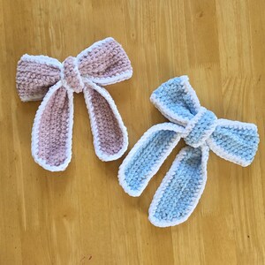 Puede incluir: Dos lazos tejidos a mano, uno en rosa empolvado y otro en azul claro, ambos con una textura suave y afelpada, con un borde de hilo blanco que perfila los bucles del lazo y las dos largas tiras que cuelgan desde el nudo central.