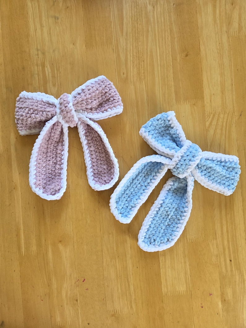 Puede incluir: Dos lazos tejidos a mano, uno en rosa empolvado y otro en azul claro, ambos con una textura suave y afelpada, con un borde de hilo blanco que perfila los bucles del lazo y las dos largas tiras que cuelgan desde el nudo central.