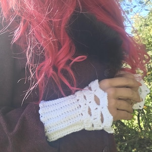 May include: A pair of white crocheted fingerless gloves featuring a ribbed texture on the wrist and a decorative openwork pattern near the knuckles, crafted from soft yarn with a scalloped edge finish at the top and bottom openings.