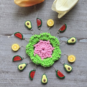 Op de afbeelding: Een handgemaakt gehaakt speldenkussen met een groen en roze bloemenpatroon. Het speldenkussen is omgeven door decoratieve spelden met fruitvormige bedels, waaronder watermeloen, avocado's en citroen.
