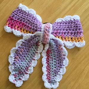 May include: A handmade crochet hair bow in shades of purple, pink, and orange, with a white scalloped edge. The bow is tied in the center with a matching crochet band, perfect for adding a touch of handmade charm to any hairstyle.
