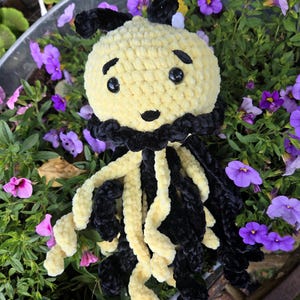 May include: A handmade crochet jellyfish plush toy featuring a round pale yellow head with black embroidered eyebrows and a small black mouth. It has two black antennae on top and long, dangling curly tentacles in alternating black and yellow yarn.