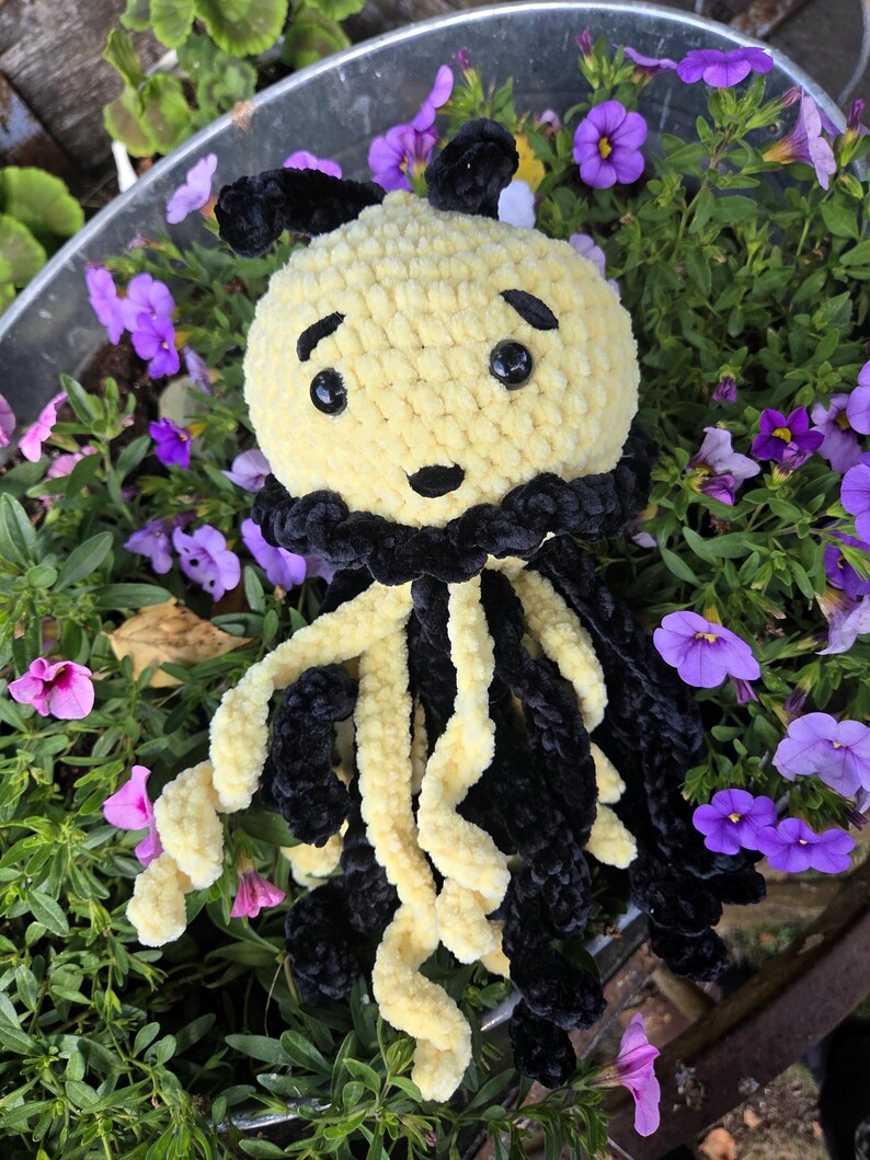 May include: A handmade crochet jellyfish plush toy featuring a round pale yellow head with black embroidered eyebrows and a small black mouth. It has two black antennae on top and long, dangling curly tentacles in alternating black and yellow yarn.