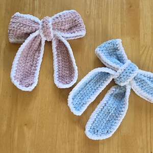 Puede incluir: Dos lazos de ganchillo hechos a mano, uno en rosa empolvado y otro en azul claro, ambos con una textura suave y afelpada con un borde de ganchillo blanco. Cada lazo tiene un nudo central y dos extremos largos que cuelgan de lana gruesa.