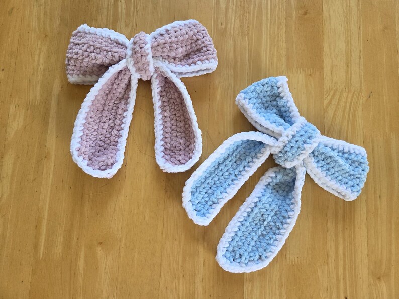 Puede incluir: Dos lazos de ganchillo hechos a mano, uno en rosa empolvado y otro en azul claro, ambos con una textura suave y afelpada con un borde de ganchillo blanco. Cada lazo tiene un nudo central y dos extremos largos que cuelgan de lana gruesa.