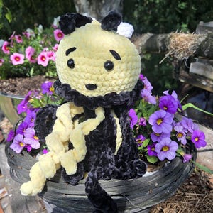 May include: A handmade crochet plush toy shaped like a bee with a round yellow head, black embroidered eyebrows, and black safety eyes. The body features alternating black and yellow yarn tentacles with a textured, soft finish and a black collar.