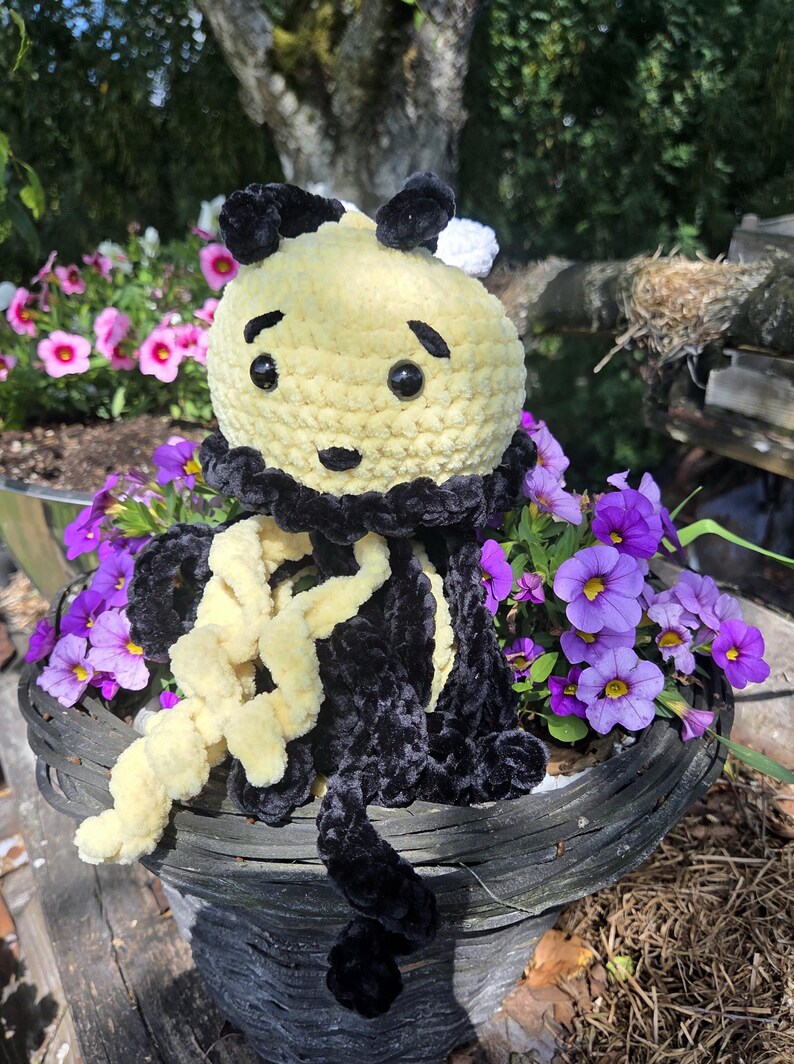May include: A handmade crochet plush toy shaped like a bee with a round yellow head, black embroidered eyebrows, and black safety eyes. The body features alternating black and yellow yarn tentacles with a textured, soft finish and a black collar.