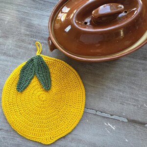 May include: A round, yellow crocheted potholder shaped like a lemon, featuring two dark green crocheted leaves at the top and a small hanging loop. Next to it sits a brown ceramic lidded casserole dish with a glossy finish.