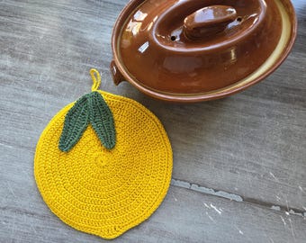 Puede incluir: Un salvamanteles redondo de ganchillo amarillo con forma de limón, que presenta dos hojas de ganchillo de color verde oscuro en la parte superior y una pequeña lazada para colgar. Al lado hay una cazuela de cerámica marrón con tapa.
