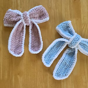 Puede incluir: Dos lazos de ganchillo hechos a mano, uno en rosa empolvado y otro en azul claro, ambos con una textura suave y afelpada con un borde de ganchillo blanco. Cada lazo tiene un nudo central y dos extremos largos colgantes de hilo grueso y suave.