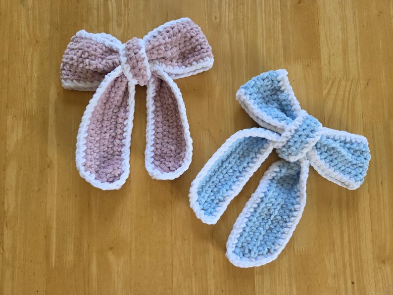 Puede incluir: Dos lazos de ganchillo hechos a mano, uno en rosa empolvado y otro en azul claro, ambos con una textura suave y afelpada con un borde de ganchillo blanco. Cada lazo tiene un nudo central y dos extremos largos colgantes de hilo grueso y suave.