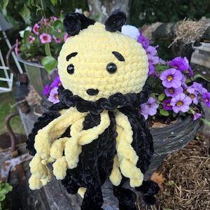 May include: A crocheted plush toy featuring a round yellow head with black embroidered eyebrows, a small black nose, and two black plastic eyes. The body is adorned with a black ruffled collar and long, dangling yellow and black curly tentacles.
