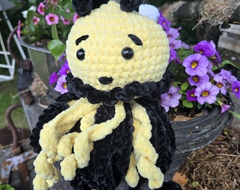 Puede incluir: Un peluche de ganchillo con una cabeza redonda amarilla, cejas negras bordadas, una pequeña nariz negra y dos ojos de plástico negro. El cuerpo presenta un cuello negro con volantes y largos tentáculos rizados de color amarillo y negro.