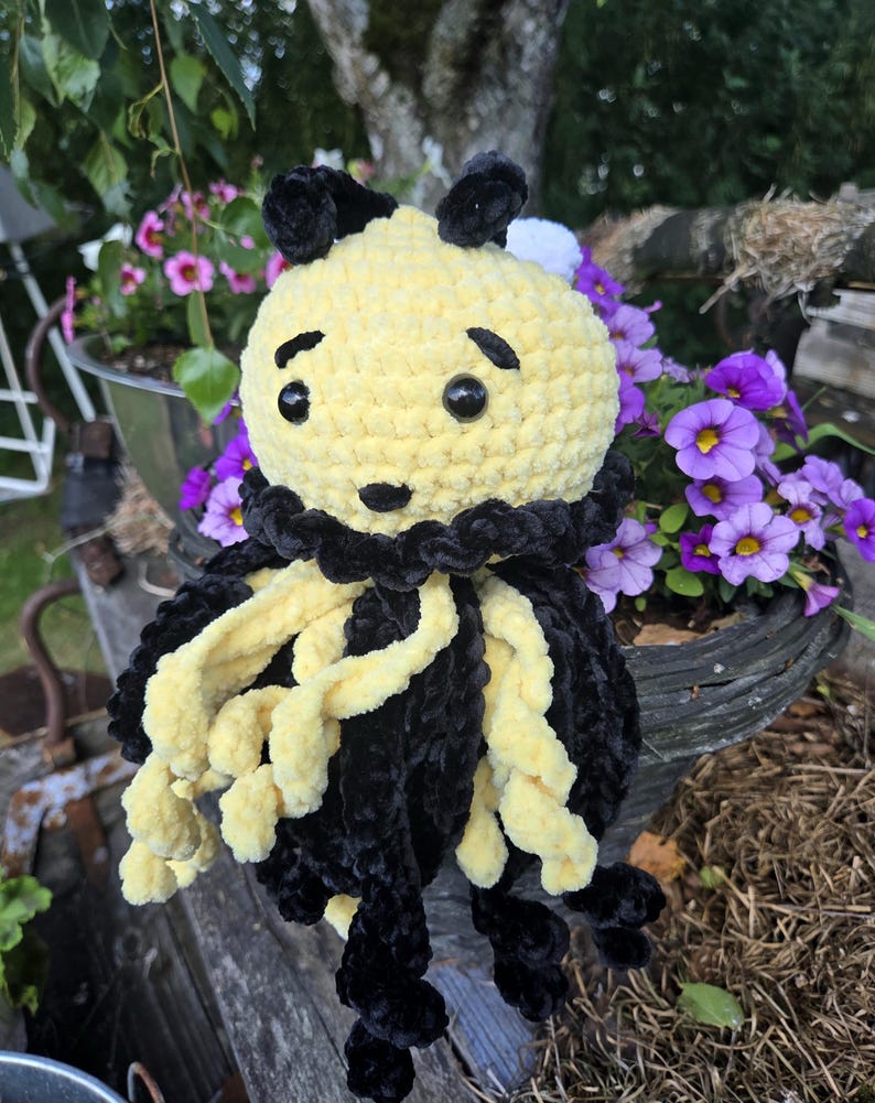 May include: A crocheted plush toy featuring a round yellow head with black embroidered eyebrows, a small black nose, and two black plastic eyes. The body is adorned with a black ruffled collar and long, dangling yellow and black curly tentacles.