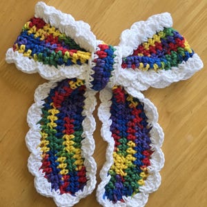 May include: A handmade crochet bow tie in a rainbow of colors, including red, yellow, green, blue, and purple. The bow tie has a white scalloped edge and is displayed on a wooden surface. The bow tie is a unique accessory.