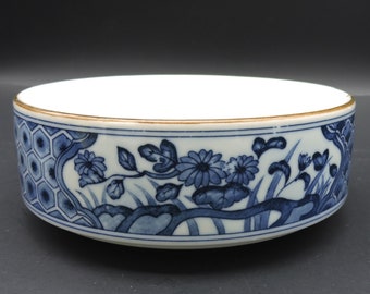 May include: A round, white ceramic bowl with a blue floral design. The bowl's exterior features a detailed pattern of flowers, leaves, and geometric shapes in shades of blue. The rim is accented with a gold band. The bowl is likely for decorative or serving purposes.