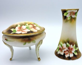 May include: A decorative porcelain trinket box and vase. The trinket box is oval, with a lid, and rests on three curved legs. The vase is tall and slender. Both items are painted with pink and white floral designs and gold trim.