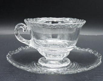 May include: A clear glass teacup and saucer set. The teacup has a decorative handle and a scalloped rim. The saucer is round with a matching scalloped edge. The set is made of clear glass and is likely vintage.
