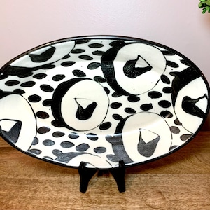 May include: Oval ceramic serving platter with a black rim and a black and white abstract design. The design features large, stylized black shapes and smaller black dots on a white background. The platter is displayed on a black stand.