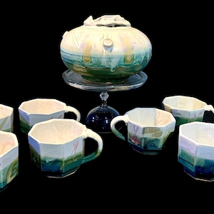 May include: A ceramic tea set consisting of a lidded teapot resting on a glass pedestal stand and seven matching octagonal mugs. Each piece features a cream-colored interior and a exterior glaze transitioning from white to green and blue hues.