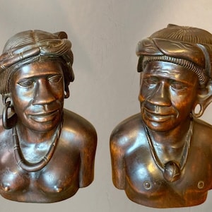 Peut inclure: Deux bustes en bois sculpté de personnes, chacun avec un visage détaillé, portant des coiffes et des colliers. Les bustes sont de couleur brun chaud, avec un grain de bois visible et des sculptures complexes. Un buste a des créoles.