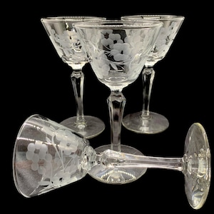 May include: A set of five clear, stemmed cocktail glasses. Each glass features an etched floral design. The glasses have a classic, elegant shape with a wide bowl and a slender stem. The glasses are arranged against a black background.