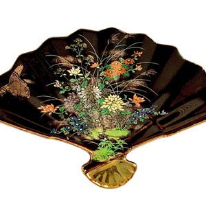 May include: A black fan-shaped decorative dish featuring a gold-trimmed edge and a detailed floral arrangement in shades of yellow, orange, green, and white. A small bird illustration is positioned on the left side of the black lacquered surface.