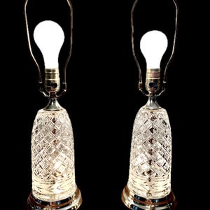 May include: Two vintage table lamps with clear, diamond-patterned glass bases and gold-toned metal accents. Each lamp features a bright white light bulb and a curved metal frame. The lamps are set against a black background.