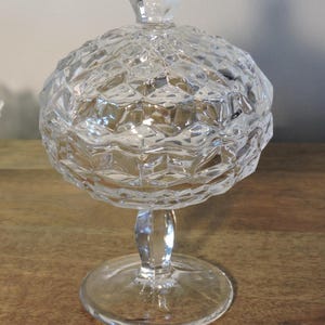 Peut inclure: Un bonbonni&egrave;re en verre transparent et facett&eacute; avec un couvercle. Le plat a une forme de bol rond et textur&eacute;, un pied et un bouton arrondi transparent. Le verre refl&egrave;te la lumi&egrave;re, cr&eacute;ant un effet scintillant. Le plat est sur une surface en bois.