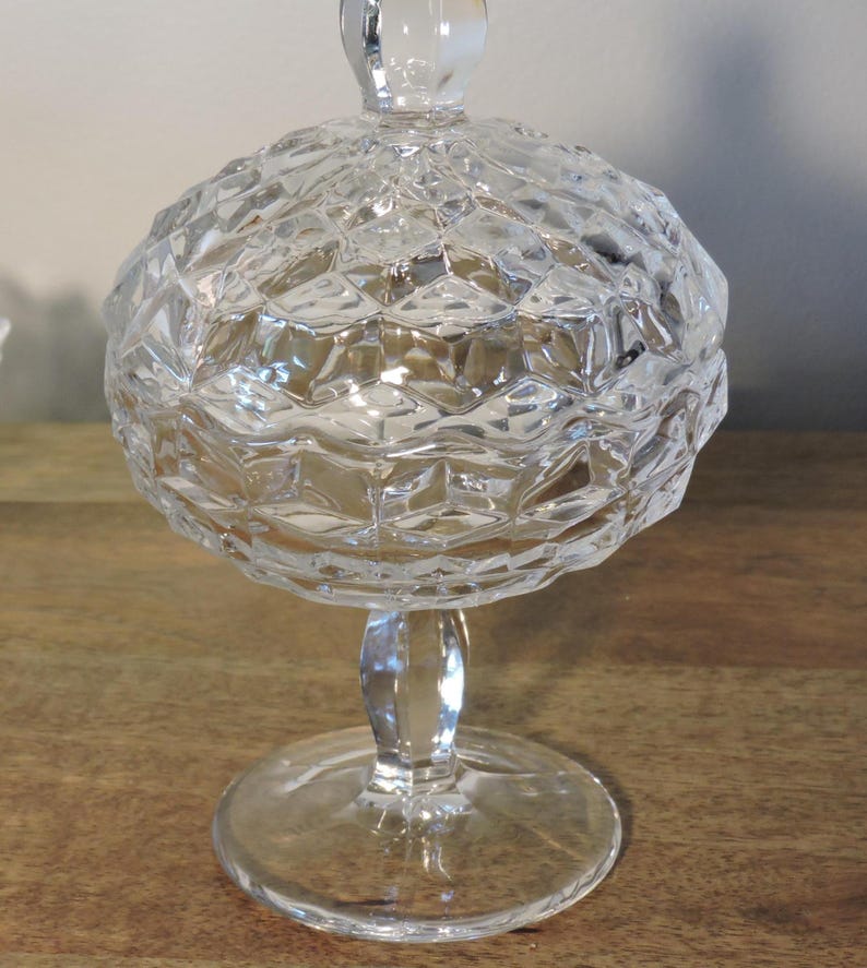 Peut inclure: Un bonbonni&egrave;re en verre transparent et facett&eacute; avec un couvercle. Le plat a une forme de bol rond et textur&eacute;, un pied et un bouton arrondi transparent. Le verre refl&egrave;te la lumi&egrave;re, cr&eacute;ant un effet scintillant. Le plat est sur une surface en bois.