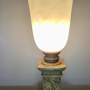 May include: A decorative table lamp with a cream-colored, tulip-shaped shade. The lamp sits on a square, column-style base with floral carvings. The base and the lamp's stem have a weathered, antique appearance. The lamp is plugged in and illuminated.