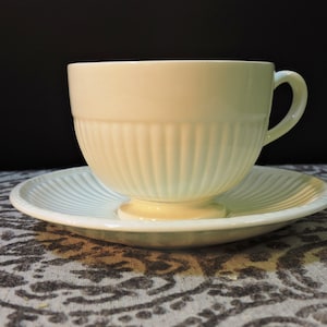 Peut inclure: Un service à thé crème composé d'une tasse et d'une soucoupe. La tasse présente des détails côtelés verticaux et une anse incurvée. La soucoupe a un motif côtelé assorti rayonnant du centre. L'ensemble est sur une surface à motifs avec un fond sombre.