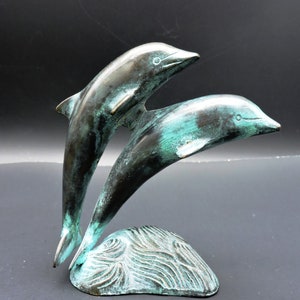 May include: Bronze sculpture of two dolphins leaping from the water. The sculpture has a verdigris patina, with shades of green and brown. The dolphins are depicted in mid-air, with their bodies arched and tails extended. The base is a stylized wave.