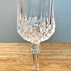 May include: A clear, cut-glass wine glass with a tulip-shaped bowl and a long, slender stem. The glass features a diamond-cut pattern on the bowl and a round base. The glass is designed for serving wine.