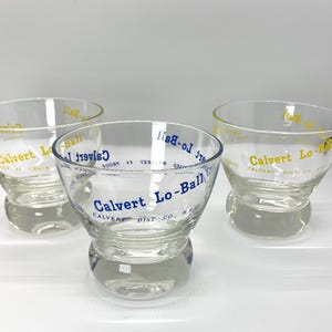 May include: Three clear glass lowball glasses with a rounded base. The glasses have a flared bowl shape and feature the text "Calvert Lo-Ball" in blue and yellow lettering. The glasses are on a white surface.