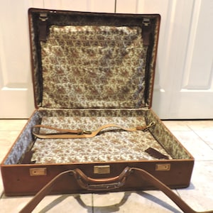 May include: Open brown vintage suitcase with a paisley patterned interior. The suitcase has a leather handle and a detachable shoulder strap. The interior features a zippered pocket and leather straps to secure items. The suitcase is open, revealing its contents.