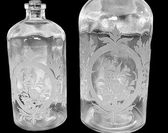 May include: Two clear glass apothecary bottles of different sizes, each topped with a cork stopper. The bottles feature an etched oval design on the front, containing a detailed floral arrangement with leaves and blossoms, set against a black background.