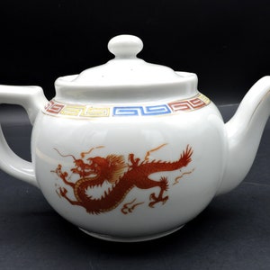 May include: White ceramic teapot with a red dragon design. The teapot has a lid and a handle. The rim is decorated with a geometric pattern in red, yellow, and blue. The spout has a gold trim.