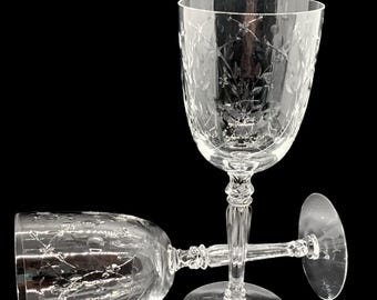 May include: Two clear crystal wine glasses with etched floral designs. One glass stands upright, while the other lies on its side. The glasses have a classic stem and base, set against a black background.