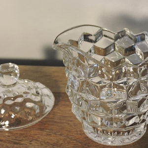 Peut inclure: Une cr&eacute;mi&egrave;re en verre transparent, facett&eacute;e, avec une anse et un couvercle assorti. La cr&eacute;mi&egrave;re et le couvercle pr&eacute;sentent un motif en forme de diamant. Le couvercle a un petit bouton rond. La cr&eacute;mi&egrave;re est sur une surface en bois.