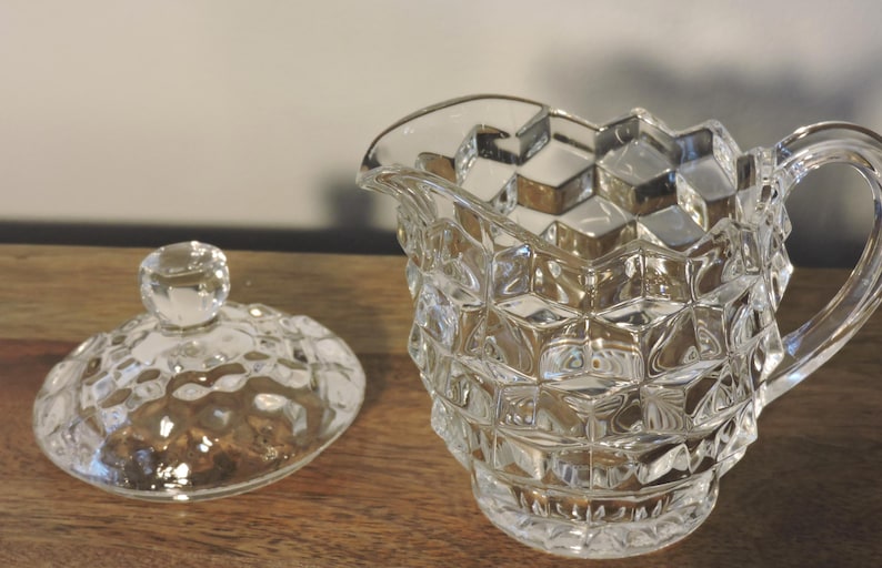 Peut inclure: Une cr&eacute;mi&egrave;re en verre transparent, facett&eacute;e, avec une anse et un couvercle assorti. La cr&eacute;mi&egrave;re et le couvercle pr&eacute;sentent un motif en forme de diamant. Le couvercle a un petit bouton rond. La cr&eacute;mi&egrave;re est sur une surface en bois.