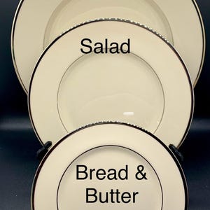 May include: A set of three ivory-colored ceramic plates with a black and silver trim. The plates are labeled "Dinner", "Salad", and "Bread & Butter". The plates are stacked on top of each other, with the largest plate at the top.